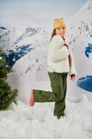 fröhliches kleines Mädchen in stylischem Outfit mit erhobenem Bein und lächelnd in die Kamera, Mode und Stil