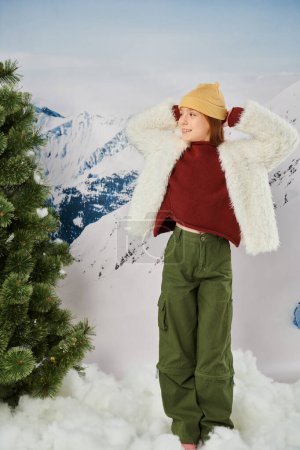 fröhliches Mädchen in stilvoller Winterkleidung posiert mit den Händen hinter dem Kopf und schaut weg, Mode