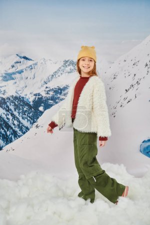 vertikale Aufnahme von preteen Mädchen in warmem Outfit mit Mütze mit schneebedeckter Bergkulisse, Mode