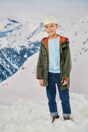 cute preadolescent boy in warm stylish attire looking at camera with mountain backdrop, fashion