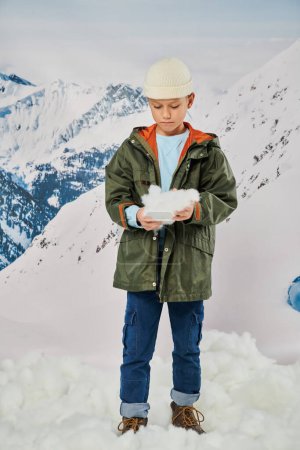 vertikale Aufnahme eines kleinen Jungen mit Schnee in den Händen im stylischen warmen Outfit, Mode