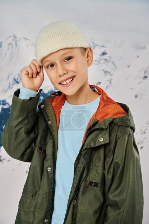Senkrechte Aufnahme eines fröhlichen kleinen Jungen in warmer Kleidung mit Hand auf Beanie-Hut, der in die Kamera lächelt