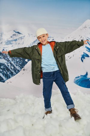 jolly preteen boy in stylish winter outfit with arms aside and smiling cheerfully at camera, fashion
