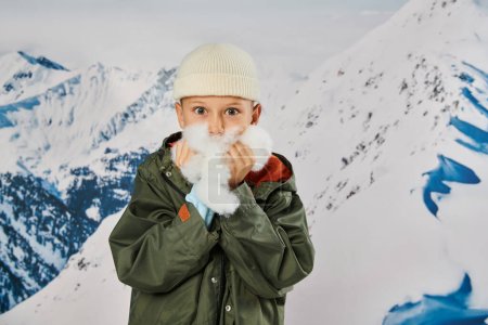 cute little boy in beanie hat posing with snow in hands near face, looking at camera, fashion