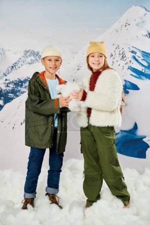vertikale Aufnahme glücklicher frühpubertärer Freunde in warmen Outfits, die mit Schnee in den Händen posieren, Mode