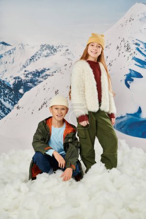 vertical shot of little boy sitting on snow near cute standing girl, stylish outfit, winter fashion