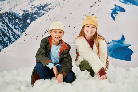 happy preadolescent boy and girl in warm attire sitting on snow and looking at camera, fashion