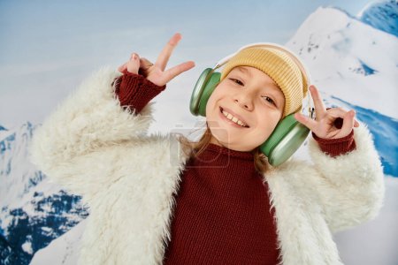 portrait of preteen girl with headset smiling joyfully at camera showing peace sign, fashion concept