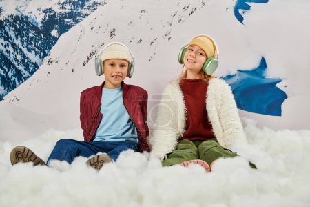 joyous trendy friends in stylish winter outfits sitting on snow with headphones, fashion concept