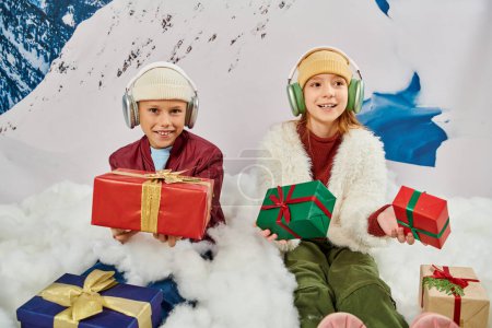 Frühchen entzückende Kinder mit Kopfhörern und Geschenke sitzen auf Schnee, lächeln in die Kamera, Mode