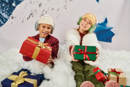 fröhliche süße Freunde in warmen Outfits mit Kopfhörern, die Geschenke halten und auf Schnee sitzen, Mode