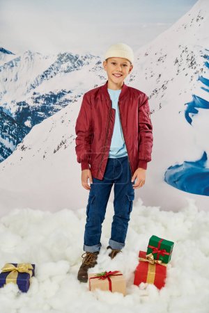 Senkrechte Aufnahme eines frühgeborenen Jungen in roter stylischer Jacke, der neben Geschenken posiert und in die Kamera lächelt