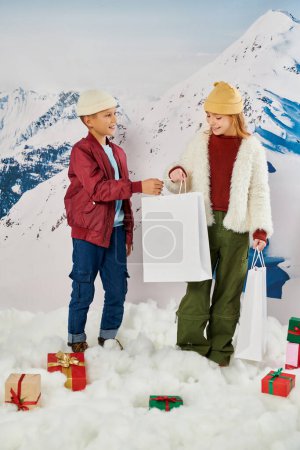 jolly preteen boy giving present bag to little girl surrounded by presents on snow, fashion concept