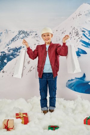 Senkrechte Aufnahme eines fröhlichen kleinen Jungen mit Geschenktüten, der in die Kamera lächelt, mit einem Berg im Hintergrund
