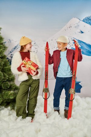 vertical shot of little girl with present next to cute boy holding skis, smiling at each other