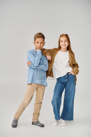 thoughtful preteen boy standing next to little jolly girl on gray backdrop, hand on shoulder, fashion