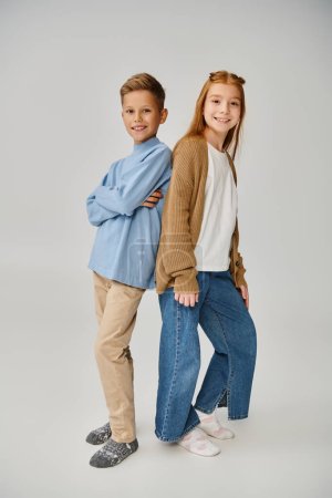 vertical shot of jolly preteen friends in casual warm outfits posing back to back on gray backdrop