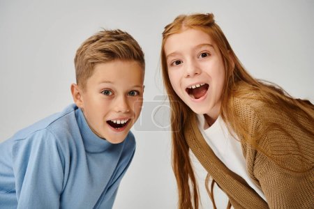 portrait of joyous little friends looking astonishingly at camera on gray backdrop, fashion concept