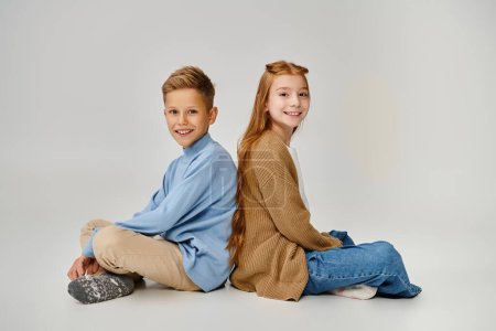 joyful preteen children sitting on floor back to back with crossed legs smiling at camera, fashion