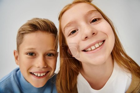 close up of two jolly preadolescent children smiling at camera on gray backdrop, fashion concept