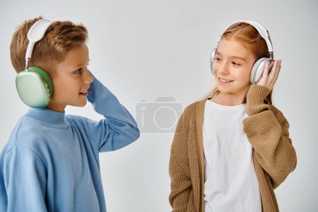 cheerful little children in warm trendy clothes with headphones smiling at each other, fashion