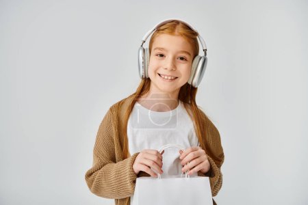 happy preteen girl in casual outfit with headset holding present bag and smiling at camera, fashion