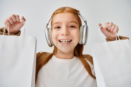 joyous little girl with modern headset posing with present bags on gray backdrop, fashion concept