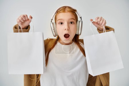 astonished little girl with headphones showing present bags at camera, Black Friday, fashion concept