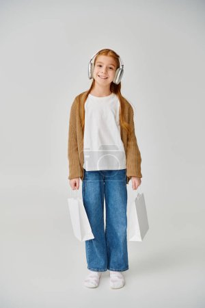 vertical shot of jolly preteen girl posing with present bags and smiling at camera, fashion concept