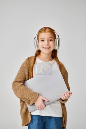 vertikale Aufnahme eines fröhlichen Mädchens mit einem Headset, das einen Laptop hält und in die Kamera schaut, Mode