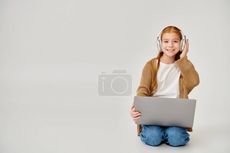 heureuse petite fille en tenue décontractée avec casque assis sur le sol avec ordinateur portable et souriant à la caméra