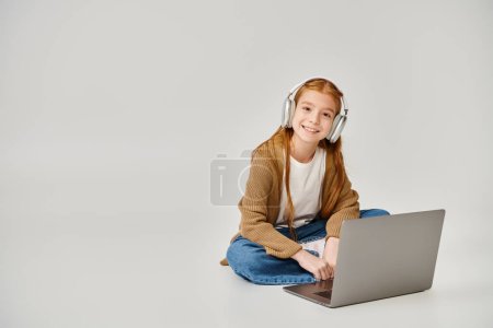 alegre chica preadolescente en el suelo con ordenador portátil y auriculares en el fondo gris, concepto de moda