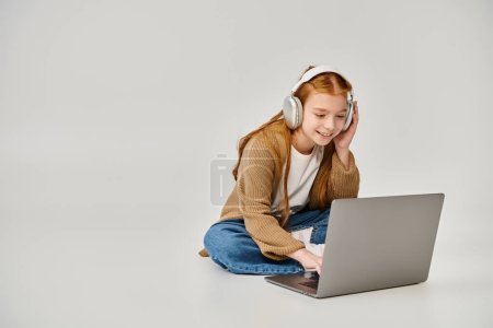 pretty cheerful little girl in winter trendy attire on floor with headset looking at laptop, fashion