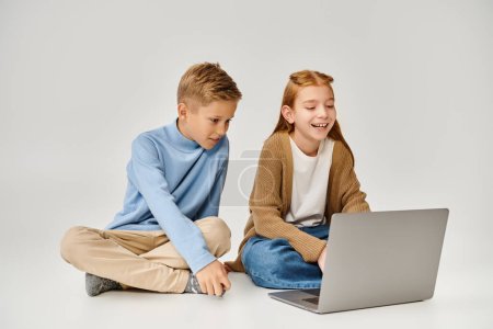 joyous preteen boy and girl sitting on floor and looking happily at laptop, fashion concept