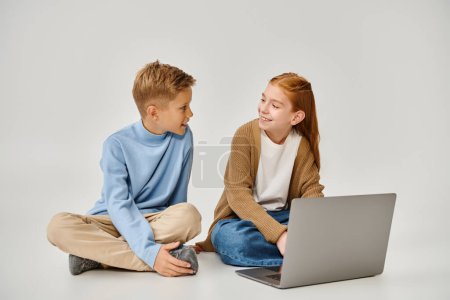 joyful preadolescent children sitting on floor with laptop and smiling at each other, fashion