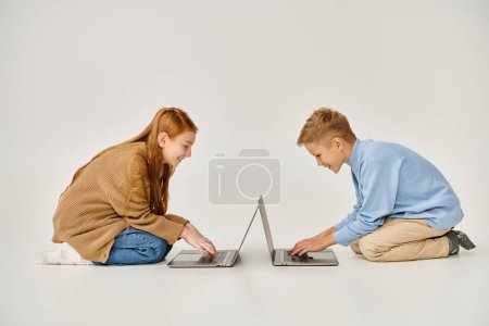 cheerful preadolescent boy and girl in trendy winter outfits sitting on floor with laptops, fashion