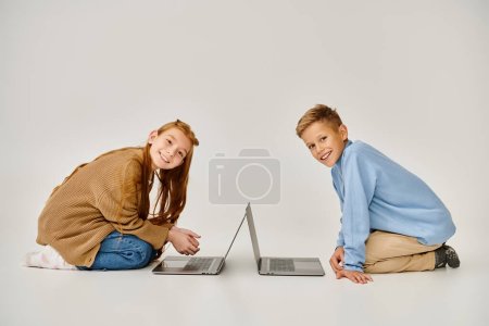 jolly preteen friends in winter clothes on floor with laptops smiling at camera, fashion concept