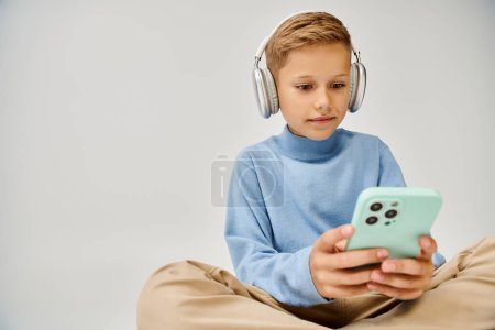 cute preadolescent boy in blue trendy sweatshirt sitting on floor with headset looking at his phone