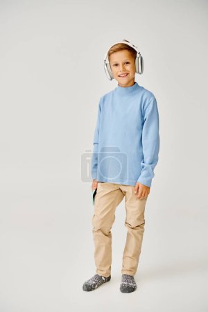 cheerful preteen boy with phone and headphones posing happily on gray backdrop, fashion concept