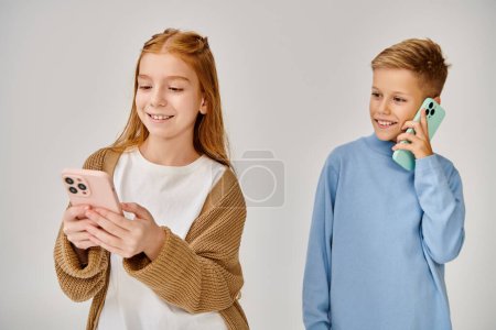 two happy preadolescent children in casual trendy attires with phones smiling joyfully, fashion