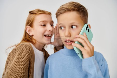 chocado niño pequeño hablando por teléfono mientras alegre preadolescente chica susurrando algo en su oído