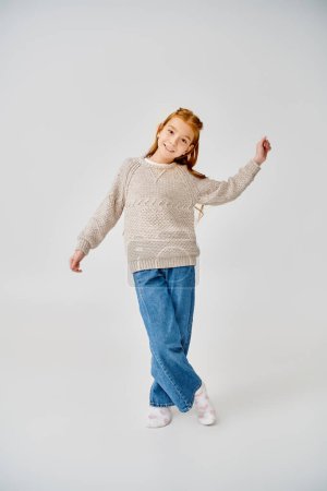 cheerful preteen girl in knitted sweater and jeans posing in motion on gray backdrop, fashion