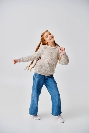 niña feliz con el pelo rojo en suéter casual posando en movimiento sobre fondo gris, concepto de moda