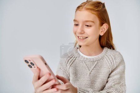 fröhliches Preteen-Mädchen mit roten Haaren, das freudig auf ihr Handy vor grauem Hintergrund blickt, Modekonzept