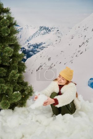 cheerful red haired little girl playing with snow next to fir tree with mountain on backdrop