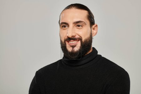 Porträt eines fröhlichen und gut aussehenden arabischen Mannes mit Bart, der in schwarzem Rollkragen vor grauem Hintergrund posiert