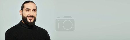 Foto de Retrato de hombre árabe alegre y guapo con barba posando en cuello alto negro sobre gris, estandarte - Imagen libre de derechos