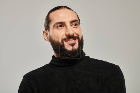 Porträt eines glücklichen und gut aussehenden arabischen Mannes mit Bart, der in schwarzem Rollkragen vor grauem Hintergrund posiert