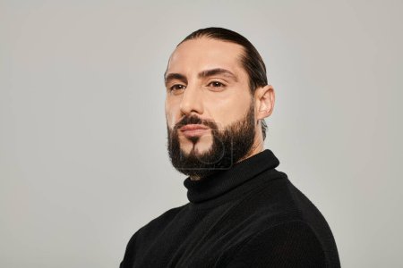 portrait of bearded and masculine arabic man posing in black turtleneck on grey background