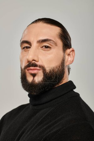 portrait of masculine arabic man with beard posing in black turtleneck on grey background